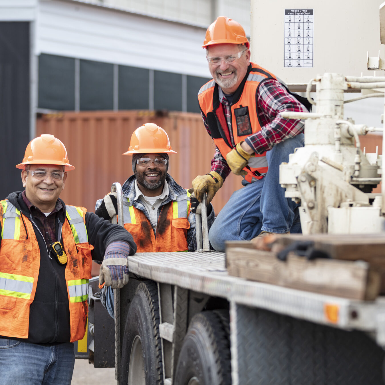 A multiracial group of three men working at a construction site, smiling at the camera. One of them is a crane operator, kneeling on the bed of his mobile crane. He is a senior man in his 60s. The other workers, standing on the ground, are mature men in their 50s.